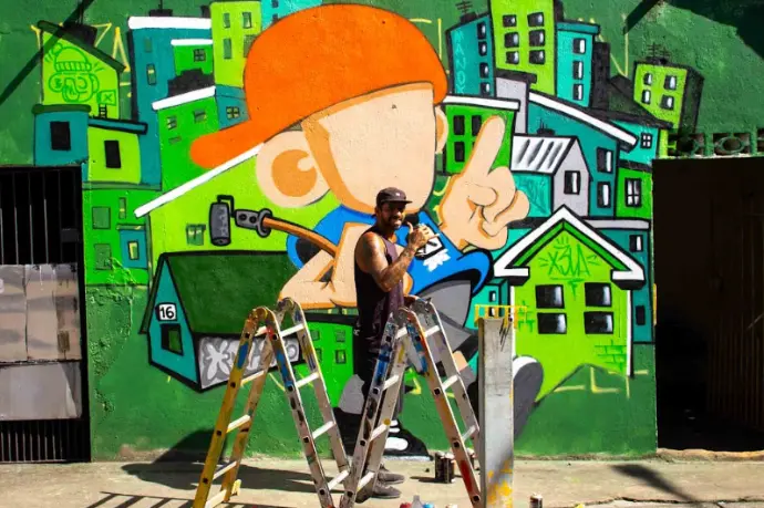 Artista posa sorridente em frente a mural colorido com personagem e cenário urbano.