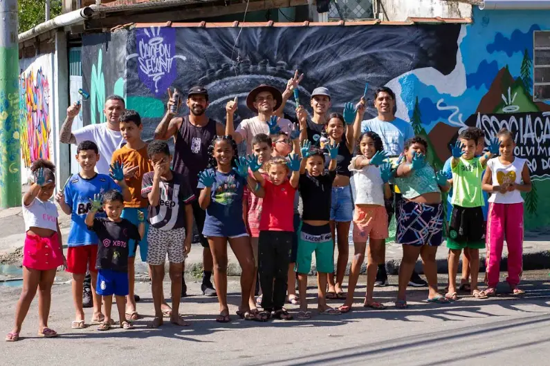 Crianças e artistas celebram em frente ao mural com mãos coloridas e muita alegria.